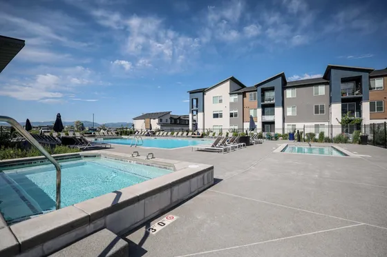 Modern apartment complex with luxurious swimming pools and stylish lounge chairs set against a picturesque backdrop of mountains and a clear blue sky.