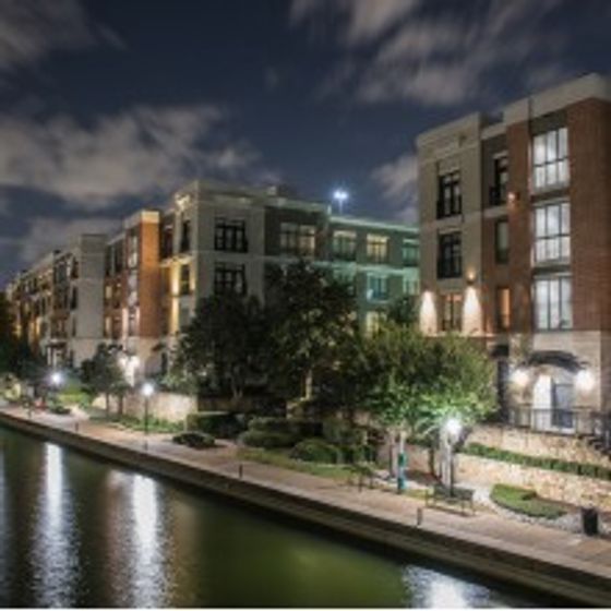 Nighttime exterior view of AMLI Las Colinas apartment building walkway along Lake Carolyn with trees and benches and lights