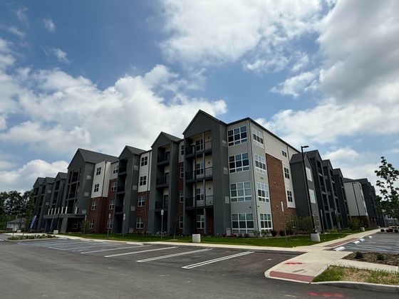 Modern apartment complex with contemporary design and ample parking, accented by lush green landscaping and a vibrant sky backdrop.