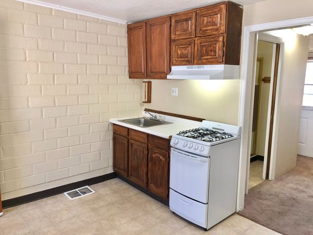 Compact kitchen featuring wooden cabinetry, gas stove, and sink area with light brick wall.