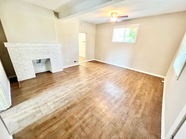 Bright living room with wooden flooring, a white brick fireplace, and ceiling fan enhancing comfort.