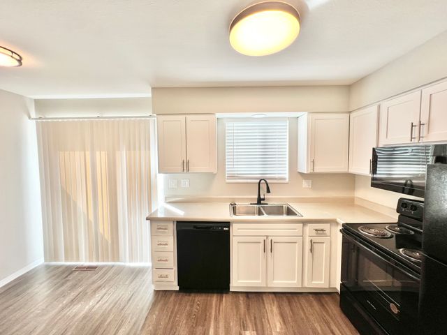 Bright modern kitchen with sleek white cabinets, black appliances, and wood flooring, featuring double sinks and ample counter space.