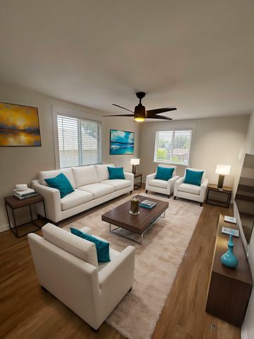 Modern living room with contemporary white seating, teal cushions, sleek wooden coffee table, and vibrant artwork, accented by a ceiling fan and natural light.
