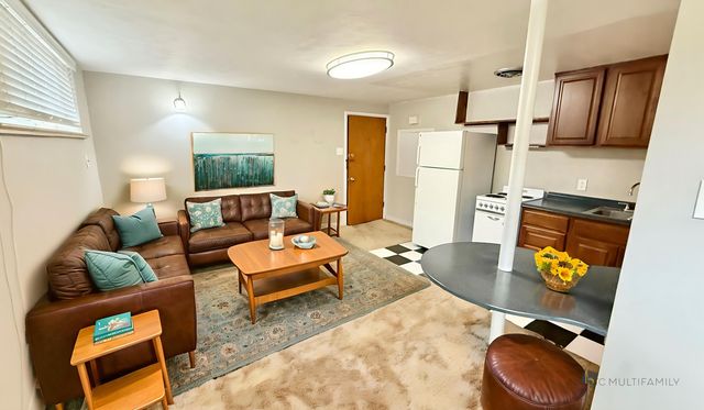 Welcoming living space featuring a cozy brown leather sectional, vibrant teal cushions, and a chic wooden coffee table, seamlessly flowing into a compact kitchen with modern cabinetry and appliances.