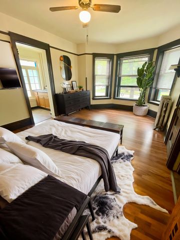 Charming bedroom with large bay windows, cozy decor, and modern accents, featuring a plush bed and vibrant indoor plant.
