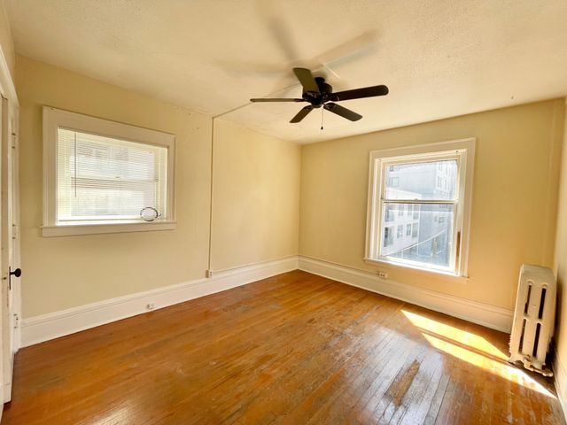 Bright and inviting empty room with polished hardwood floors, two sunlit windows, and a classic ceiling fan.