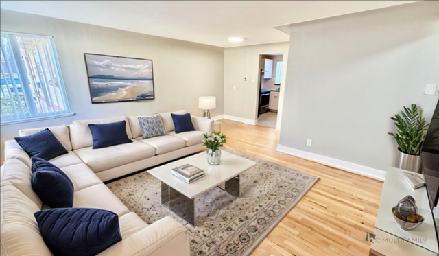 Bright and modern living room with elegant light wood flooring, large white L-shaped sofa, decorative cushions, and a sleek coffee table, accentuated by natural light from the window.