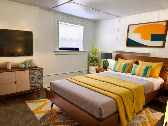 Chic bedroom with modern decor, featuring a stylish bed adorned with vibrant pillows and a cozy mustard blanket. Includes a sleek TV, elegant artwork, and a lush indoor plant for a touch of nature.