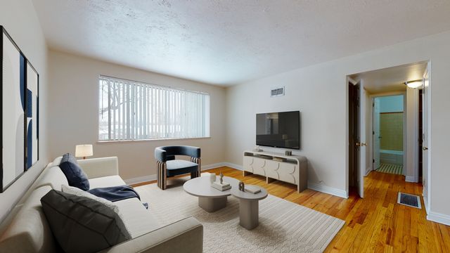 Bright and inviting living room with modern decor, featuring a comfortable sofa, stylish coffee tables, and a large flat-screen TV; natural light streaming through a window with vertical blinds enhances the warm wood flooring.
