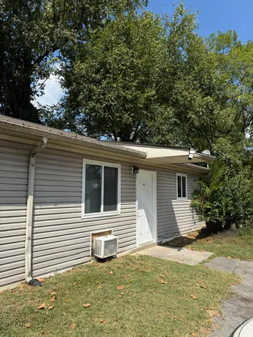 Charming single-story home with modern siding, surrounded by lush green trees and a well-maintained lawn, inviting entrance under a sunny blue sky.