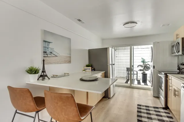 Modern kitchen with sleek countertops, stylish stools, and a well-lit dining area leading to a patio with potted plants and outdoor seating.