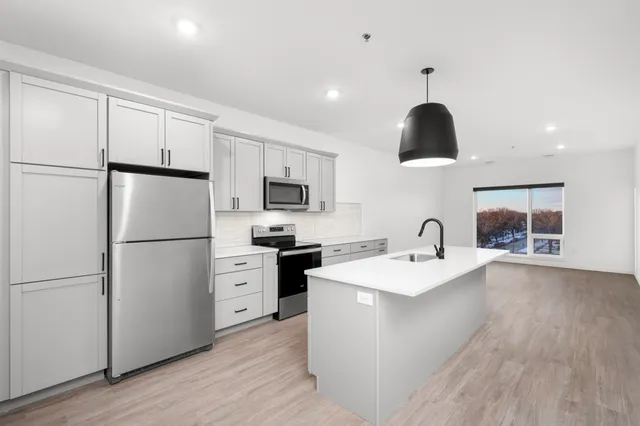 Modern open-concept kitchen with sleek gray cabinets, stainless steel appliances, and a stylish island overlooking a bright living area with large windows.