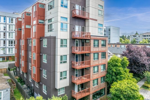 Modern multi-story apartment building with vibrant red balconies, surrounded by lush green trees, under a clear blue sky.