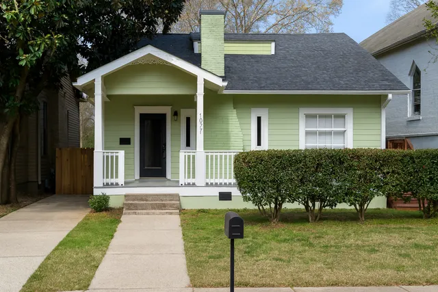 Charming single-story green home with white trim, cozy front porch, and manicured hedges in a friendly neighborhood setting.