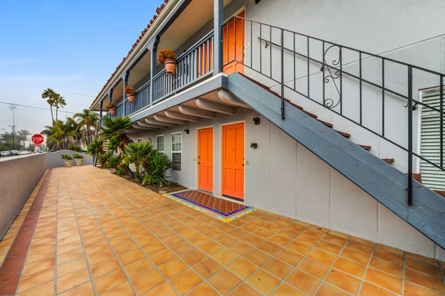 Charming apartment exterior with vibrant orange doors, stylish wrought iron railings, and lush palm plants on a spacious tiled patio.