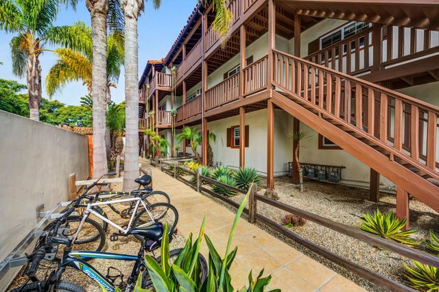 Charming apartment complex with rustic wooden balconies and lush landscaping, featuring bike parking and palm trees under a clear blue sky.