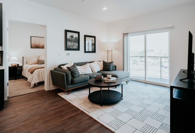 Bright, modern living room with cozy gray sofa, stylish coffee table, and inviting bedroom view through open door, enhanced by natural light from sliding glass doors.