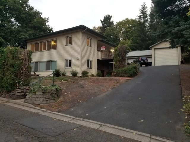 Charming two-story house with lush greenery, large windows, and a spacious driveway leading to a detached garage, ideal for family living.