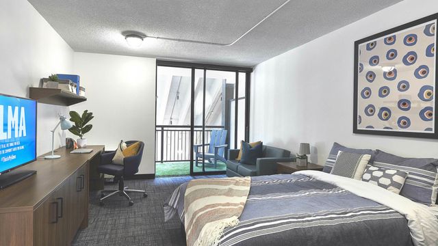 Modern bedroom featuring a cozy bed with stylish pillows, a sleek desk with a chair, and a balcony view through floor-to-ceiling glass doors.