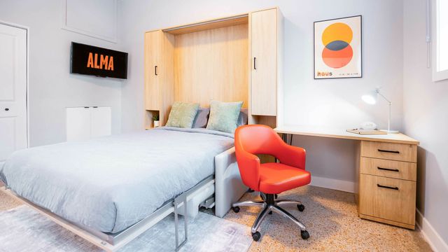 Bright modern bedroom with a wall-mounted TV, cozy bed with wooden frame, and stylish workspace featuring a vibrant red chair and sleek desk.