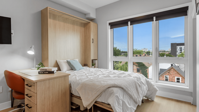 Bright bedroom with large windows, cozy bed, and stylish wooden desk; perfect for a serene home office setup.