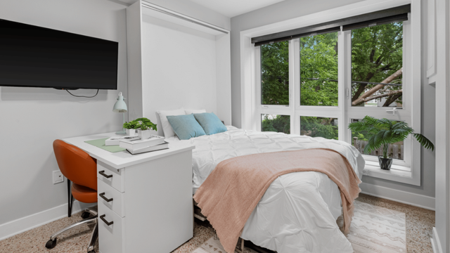 Bright bedroom with large windows, cozy bed with pastel pillows, and a sleek desk with modern decor.