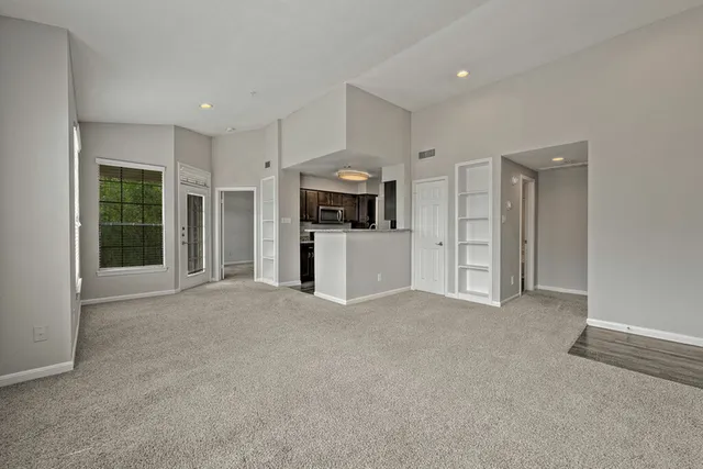 Bright, open-concept living room with plush carpet, tall ceilings, and modern kitchen access.