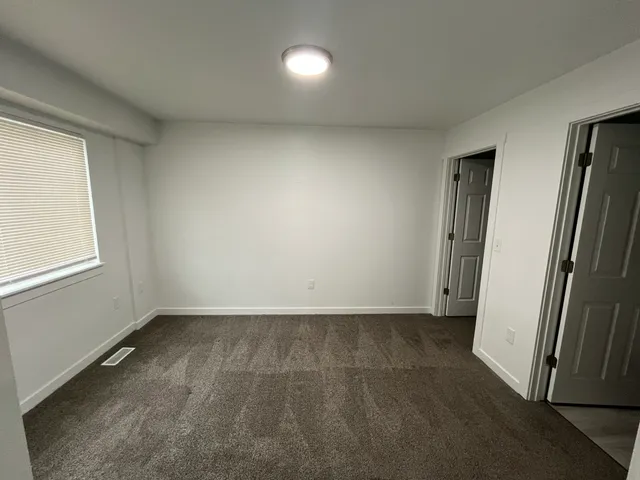 Bright and spacious room with clean carpet, fresh white walls, and ample natural light from a large window, featuring two doors for versatile access.