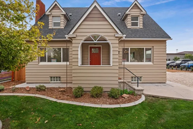 Charming two-story beige house with a red front door, dormer windows, and a neatly landscaped front yard, set against a clear blue sky backdrop.
