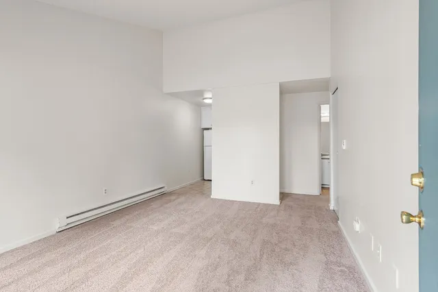 Bright and spacious living area with plush carpet and high ceiling, offering an open view into a cozy kitchen with a white refrigerator.