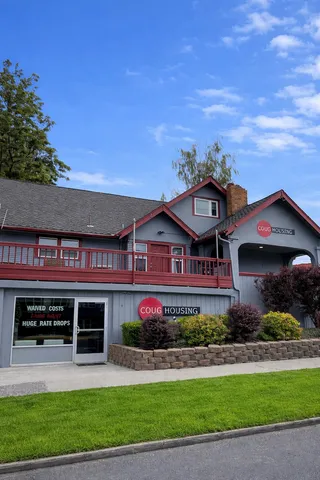 Charming gray and red two-story building with lush landscaping and bright blue sky, featuring Coug Housing signage and promotional offers.