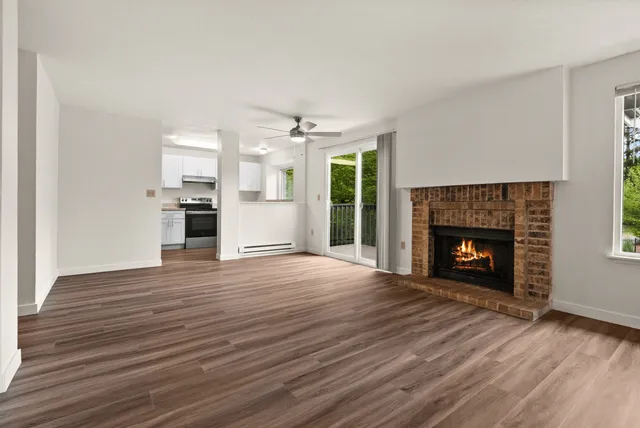 Open-concept apartment interior featuring a cozy brick fireplace, modern wood-style flooring, and a view into a bright white kitchen with stainless steel appliances.