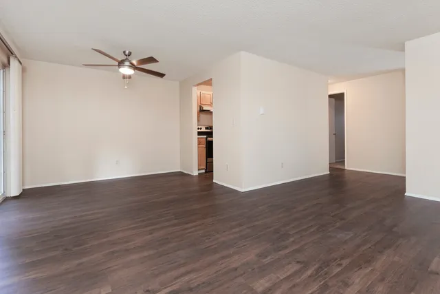 Spacious empty living room with dark wood flooring, beige walls, a ceiling fan, and an open view into the kitchen.
