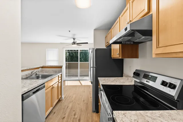 Modern kitchen with sleek stainless steel appliances, marble countertops, and ample cabinetry, leading to a bright living area with wooden floors and a ceiling fan.