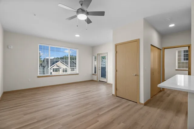 Bright and spacious living area with modern ceiling fan, large window showcasing beautiful residential view, laminate flooring, and access to an outdoor balcony.