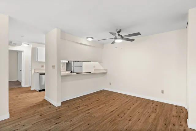 Bright open-plan living area with modern ceiling fan and hardwood floors leading to a sleek kitchen with stainless steel appliances.