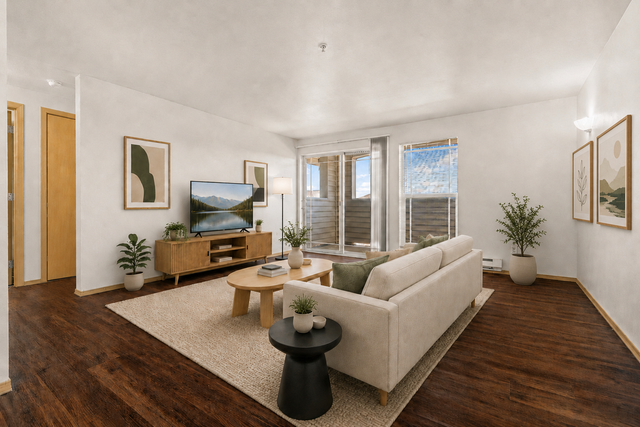 Inviting living room with a cozy beige sofa, elegant wooden flooring, modern art pieces, and a TV on a stylish console, bathed in natural light from large windows.