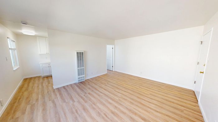 Bright and airy room with sleek wooden floors and clean white walls, leading into a cozy kitchen with modern cabinetry.