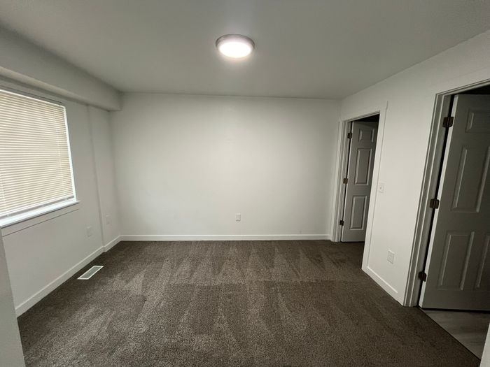 Bright and spacious room with clean carpet, fresh white walls, and ample natural light from a large window, featuring two doors for versatile access.