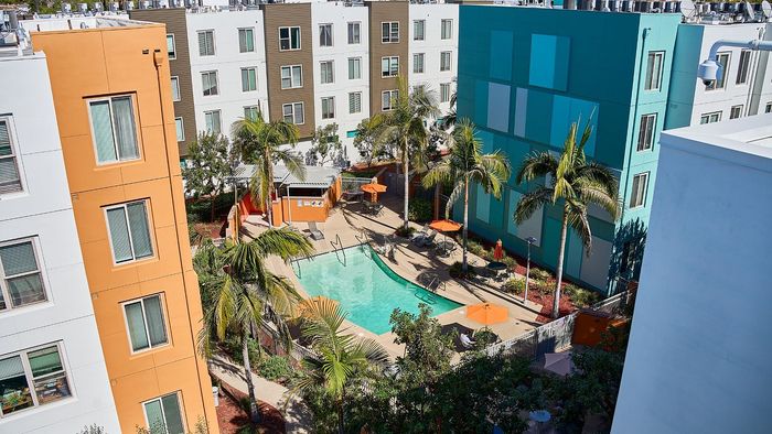 Vibrant apartment complex featuring a sparkling pool, surrounded by modern colorful buildings, palm trees, and relaxing outdoor seating.