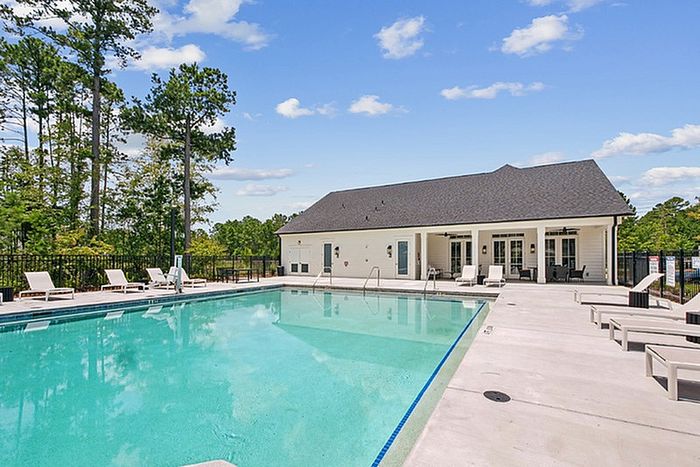 Inviting poolside view with sleek sun loungers and a charming clubhouse nestled amidst lush greenery and clear blue skies.