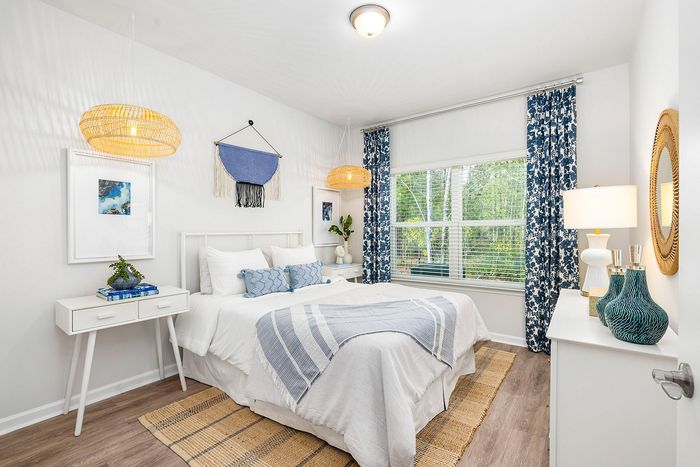 Bright and airy bedroom with chic decor, featuring a cozy bed adorned with blue accents, stylish woven lamps, and large windows offering lush garden views.