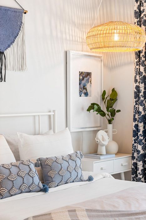 Charming bedroom with elegant decor featuring blue and white patterned pillows, stylish wicker pendant light, and vibrant greenery. A cozy ambiance with artistic wall hangings and a sleek nightstand completes the serene look.
