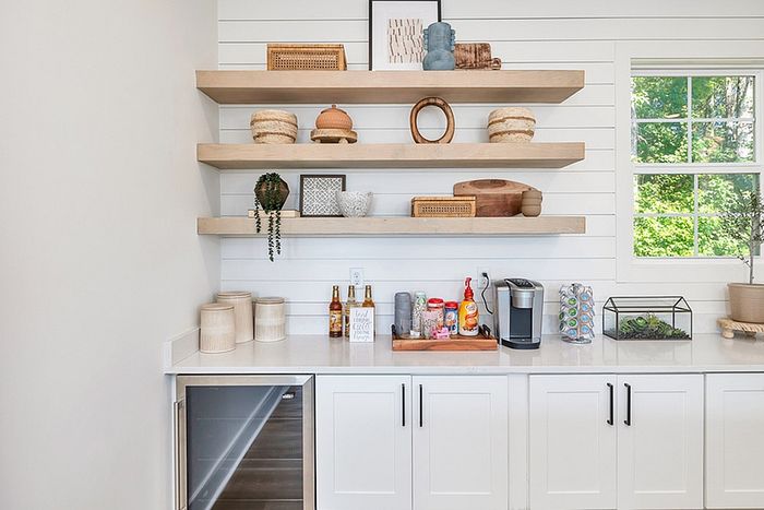 Chic kitchen nook with floating shelves displaying decorative items, well-lit counter space, and a window view of greenery.