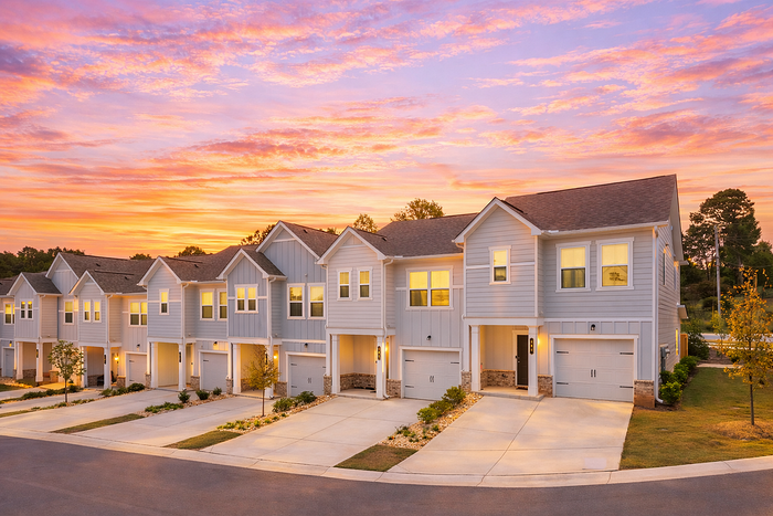 Charming row of modern townhouses with light blue siding, warmly lit windows, and beautifully landscaped lawns, set against a stunning sunset sky.