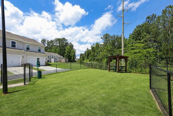 Charming townhouse community with lush green lawn, scenic wooded backdrop, and private fenced area under a clear blue sky.