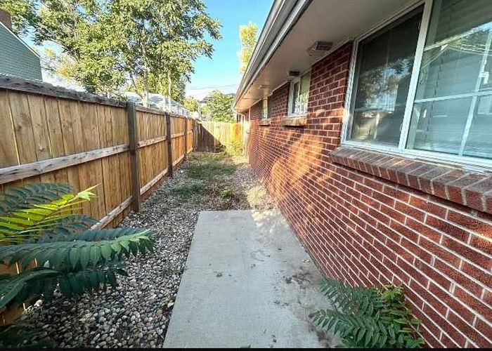 Inviting side walkway with brick exterior and wooden fence, featuring greenery and a clean path.