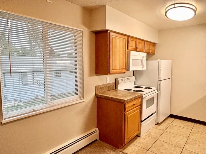 Cozy kitchen in modern apartment featuring large window with blinds, wooden cabinets, and sleek white appliances.
