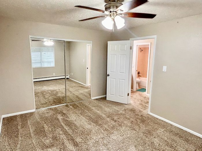 Bright and spacious bedroom featuring plush carpet, a mirrored closet, and modern ceiling fan beside an ensuite bathroom.