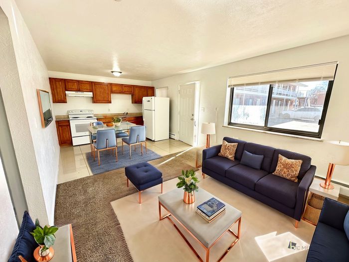 Bright and inviting living room with a modern kitchen and dining area, featuring a cozy navy sofa, stylish copper accents, and large windows for abundant natural light.